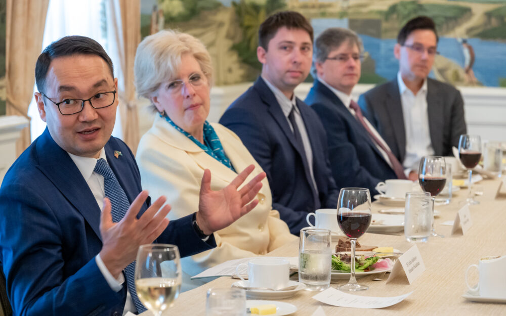 A photo from a luncheon shows HE Yerzhan Ashikbayev, Ambassador of Kazakhstan, gesturing with his hands while speaking. He is seated at a table with other attendees, including Nathan Depew, who is visible in the center of the image. Glasses of wine and water are on the table.